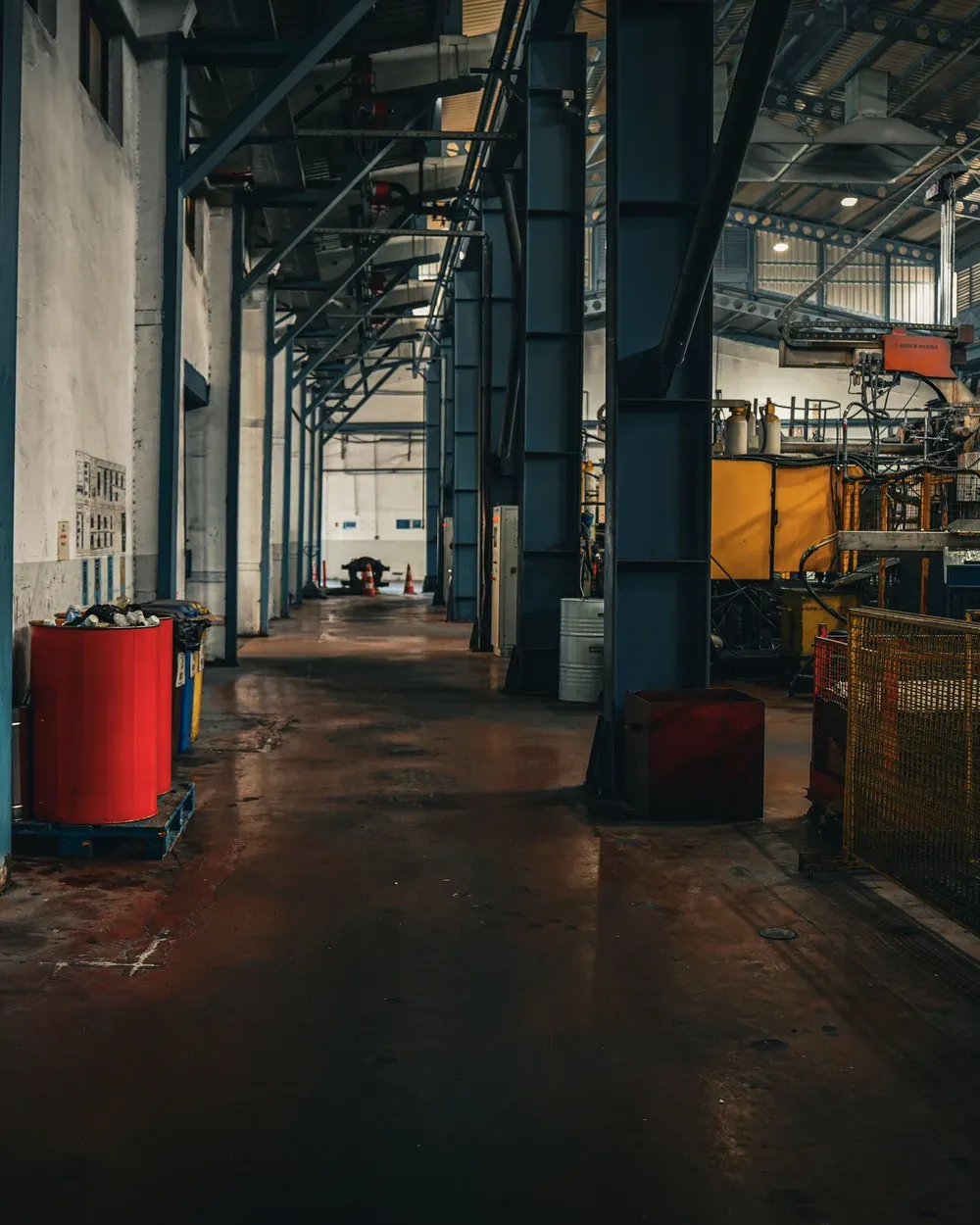 Corredor de galpão industrial, com estrutura metálica azul, maquinário ao fundo e piso de concreto molhado