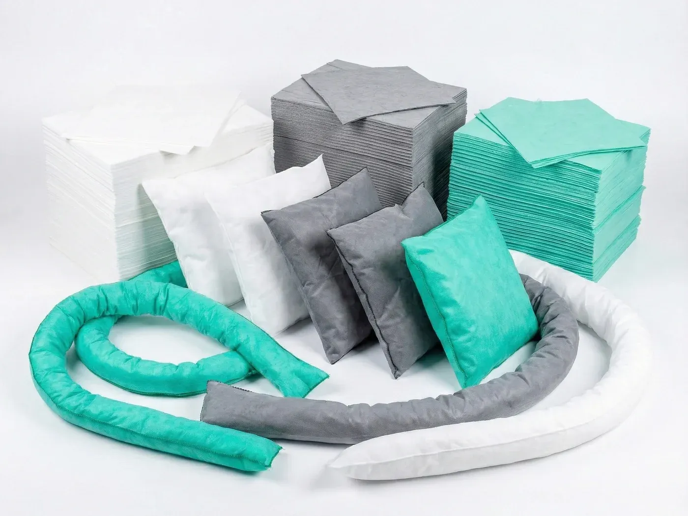 A linha completa reunida: pilhas de mantas branca, cinza e verde ao fundo, travesseiros das três cores ao centro e cordões em laço à frente