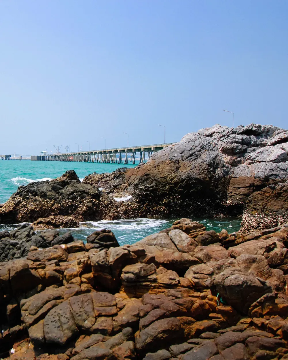 Costa rochosa com o mar turquesa quebrando entre as pedras e um píer industrial ao longe