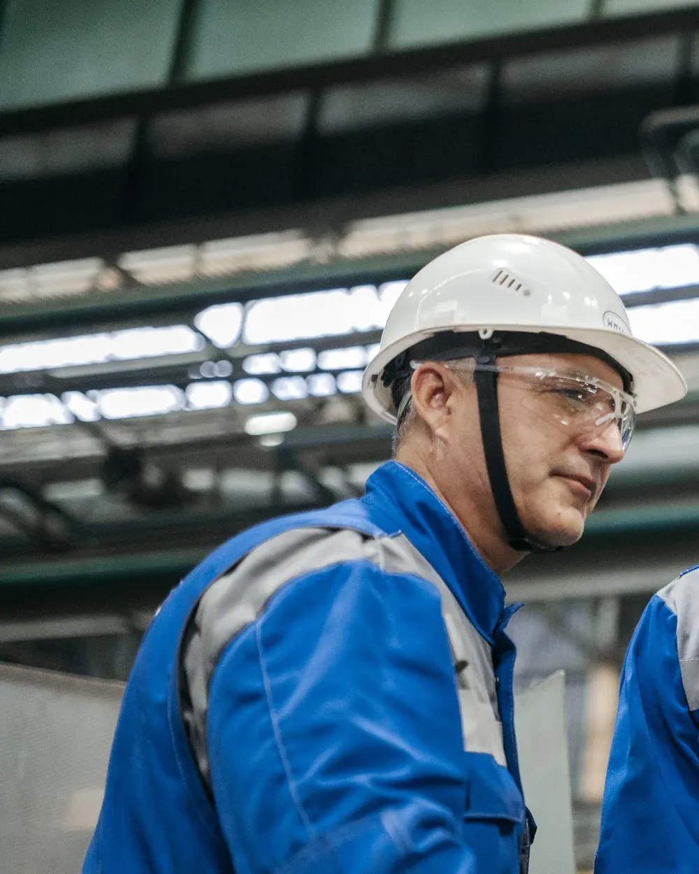 Operador de uniforme azul, capacete branco e óculos de proteção, de perfil, numa planta industrial